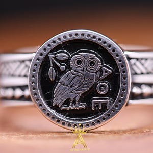 May include: A silver ring featuring a black enamel owl design. The owl is encircled by a ring of small dots and the band of the ring has a textured pattern.