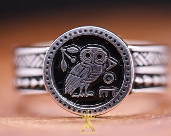 May include: A silver ring with a black enamel owl design. The owl is surrounded by a circle of small dots and the ring band has a textured design.