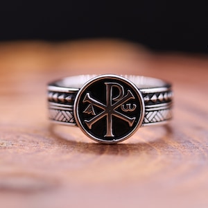 Può includere: Un anello d'argento con un centro in smalto nero che presenta un simbolo cristiano stilizzato, il Chi Rho, con le lettere 'P' e 'X' che si incrociano. L'anello ha una fascia testurizzata con un motivo geometrico.