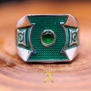 Puede incluir: Un anillo de tono plateado con una cara rectangular que presenta una incrustación de esmalte verde texturizado y una piedra verde circular central. Los lados del anillo muestran un emblema geométrico verde. El anillo incluye el texto SILVER MASTER y un logo de corona.