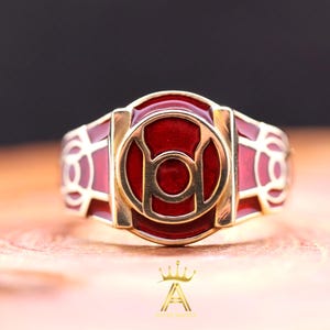Op de afbeelding: Een goudkleurige ring met een rood emaille ontwerp. De ring heeft een centraal cirkelvormig embleem met een kleinere cirkel binnenin, omgeven door een gouden rand. De band van de ring heeft een herhalend geometrisch patroon.