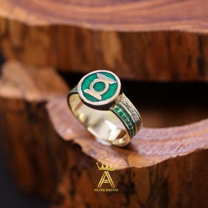 May include: A gold ring with a green enamel emblem featuring a white design. The ring has a detailed band with green accents. The ring is displayed on a wooden surface.