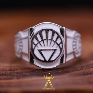 May include: A white metal ring featuring a white enamel design of a stylised sun with a triangle inside. The ring is displayed on a brown wooden surface.