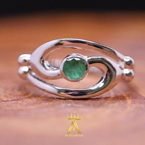 May include: A silver ring featuring a central round, faceted green gemstone set within a polished, swirling metal band. The design includes two small spherical accents on each side of the band. A gold crown logo with the text SILVER MASTER is visible.