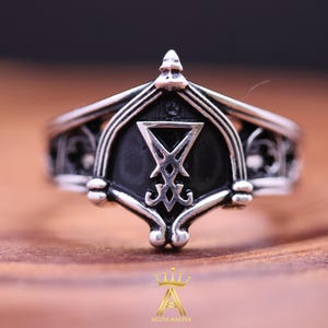 Könnte beinhalten: Ein silberner Ring mit einem schwarzen Emblem, das ein stilisiertes umgekehrtes Dreieck und andere Symbole zeigt. Der Ring hat ein verziertes Design mit gotischen Bögen und dekorativen Elementen. Der Ring liegt auf einer Holzoberfläche.