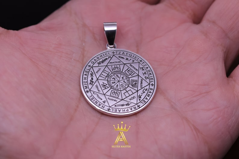 May include: Silver pendant featuring a circular design with a seven-pointed star in the center. The star is surrounded by a circle with the names of angels written in Hebrew and English, including Michael, Gabriel, Raphael, and Samael.