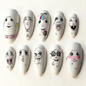 May include: A set of ten white almond-shaped press-on nails, each featuring a unique hand-drawn cartoon character face with distinct expressions, including a lollipop, glasses, and blushing cheeks, rendered in black ink with subtle pink and color accents.