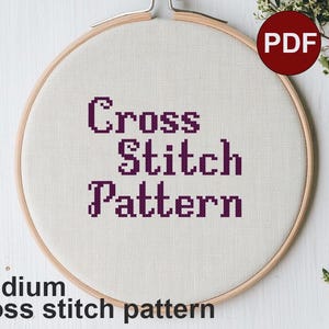 May include: A close-up of a white embroidery hoop with the words "Cross Stitch Pattern" in purple text. The hoop is on a white wooden surface.