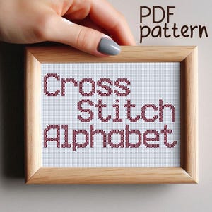 May include: A wooden frame with a white background and the text "Cross Stitch Alphabet" in red cross stitch.