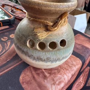 May include: A round ceramic vessel with a wide, flared rim and a textured, mottled finish in shades of beige, brown, and muted green. A thick, braided jute rope is tied around the neck, and a row of four circular cutouts encircles the upper body.