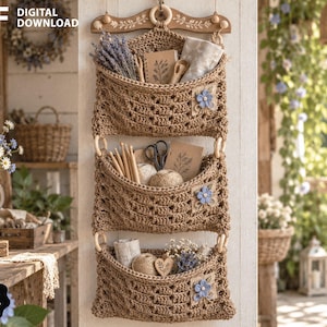 Puede incluir: Un organizador de almacenamiento colgante de tres niveles hecho de yute tejido en color canela, suspendido de un colgador de madera decorativo. Cada uno de los tres bolsillos abiertos tiene una pequeña flor de tela azul y contiene diversos materiales.