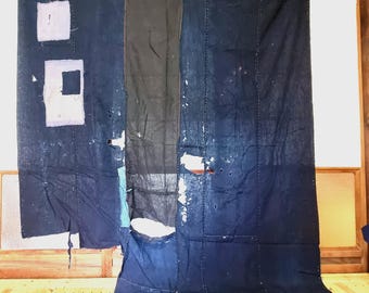 May include: A large, indigo-dyed textile with visible stitching and patches. The fabric features a central black panel and several white and light blue patches. The textile is displayed against a wooden frame and floor.
