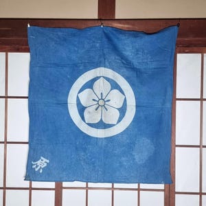 Könnte beinhalten: Blaues Stoffbanner mit einem weißen Blumenemblem in einem Kreis. Das Banner hat ein verblasstes Aussehen und ein japanisches Zeichen unten links. Es hängt vor einem Shoji-Bildschirm mit Holzrahmen.