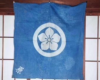 May include: Blue fabric banner with a white floral emblem inside a circle. The banner has a faded appearance and a Japanese character in the lower left corner. The banner is hanging in front of a wooden framed shoji screen.
