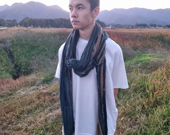 May include: A young person wearing a white t-shirt and a blue and brown striped scarf. The person is standing in a field with mountains in the background.