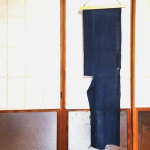 以下が含まれることがあります： 木製の棒から吊るされた、長い濃い青色の織物。この生地は、着物または同様のアイテムである可能性のある、伝統的な日本の衣服のようです。背景には、伝統的な日本の障子スクリーンがあります。