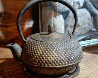 May include: A small, antique cast iron teapot with a textured, dimpled surface and a curved handle. The teapot has a spout and a lid with a small knob. It sits on a matching circular base, showcasing a rustic, aged appearance.