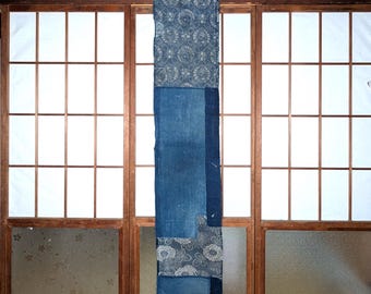 May include: A long, vertically-oriented textile with a patchwork design, featuring various shades of indigo blue. The top section has a floral pattern, while the lower sections showcase solid and patterned fabrics. The textile hangs in front of a wooden-framed window.