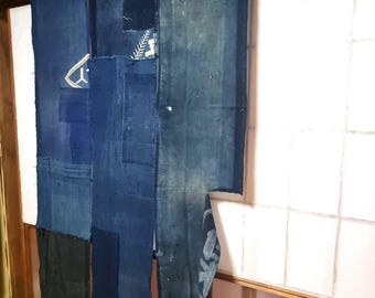 May include: A patchwork textile curtain in shades of indigo blue, with visible stitching and varying fabric textures. Patches of darker and lighter blue create a visual contrast. The curtain hangs in a doorway, with a glimpse of a white shoji screen.