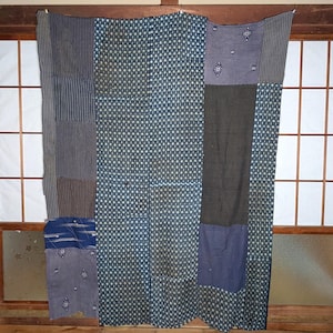 Puede incluir: Textil de patchwork con una variedad de telas azules y grises. El diseño incluye rayas, pequeños patrones geométricos y secciones lisas. El textil está colgado frente a una ventana con marco de madera.