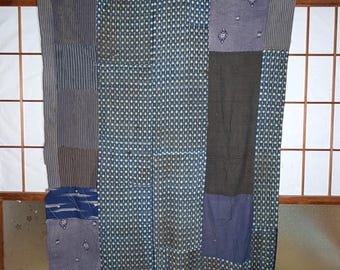 May include: A patchwork textile with a variety of blue and gray fabrics. The design includes stripes, small geometric patterns, and solid sections. The textile is hanging in front of a window with a wooden frame.