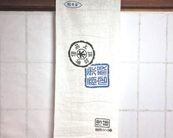 May include: A cream-colored linen hanging scroll with Japanese text in blue and black ink. The scroll features several circular and rectangular stamps with intricate characters. The top of the scroll has a small oval stamp.