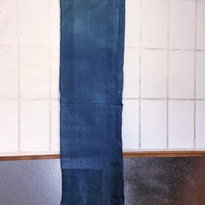 Könnte beinhalten: Ein rechteckiges, indigogefärbtes Textil, das an einer Holzstange hängt. Der Stoff zeigt einen Farbverlauf von Blautönen, wobei die Farbtöne am unteren Ende dunkler sind. Es ist wahrscheinlich ein Wandbehang oder ein dekoratives Element.