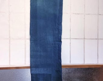 May include: A long, rectangular indigo-dyed textile hanging from a wooden rod. The fabric displays a gradient of blue hues, with darker shades at the bottom. The textile is likely a wall hanging or decorative piece.