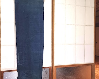 May include: A long, indigo-dyed textile hanging from a wooden rod. The fabric has visible patches and a faded appearance, suggesting age. The background features a traditional Japanese shoji screen with white paper panels and wooden frames.
