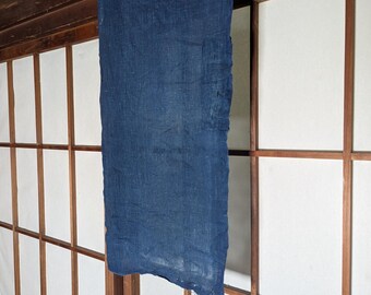 May include: A rectangular piece of indigo-dyed fabric hangs vertically. The fabric has a textured appearance and is suspended in front of a traditional Japanese shoji screen with wooden frames and white paper panels. The fabric's edges are uneven.