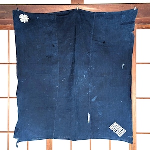 May include: A rectangular indigo-dyed textile featuring a deep blue hue with visible variations in colour intensity. The fabric displays a white embroidered flower in the upper left corner and a white geometric kanji character in the lower right.