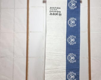 May include: A rectangular, vertically-oriented textile banner with a wooden hanging rod. The banner is divided into two vertical sections: a white section and a blue section with white floral designs. Japanese text is printed on the white section.