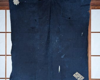 May include: A dark indigo textile hanging with a white floral applique in the upper left corner. The textile has visible seams and some wear and tear. A Japanese character design is in the lower right corner.