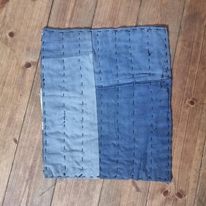 Könnte beinhalten: Quadratischer Patchwork-Textil in Blautönen mit sichtbaren Nähten. Der Stoff ist in zwei vertikale Abschnitte mit unterschiedlichen Blautönen unterteilt. Das Stück liegt auf einer Holzoberfläche und zeigt sein handwerkliches Aussehen.