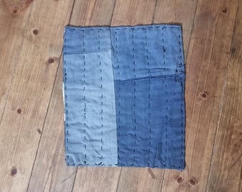 May include: A square, patchwork textile in shades of blue, with visible stitching. The fabric is divided into two vertical sections of varying blue hues. The piece is placed on a wooden surface, showcasing its handcrafted appearance.