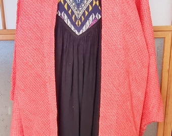 May include: A coral pink knitted cardigan with a geometric patterned black dress underneath. The cardigan has a shawl collar and long sleeves.