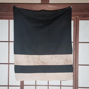 Könnte beinhalten: Ein schwarz-beige gestreifter Stoff hängt vor einem Holzrahmen und einem Shoji-Bildschirm. Der obere Teil ist einfarbig schwarz, mit zwei beigen Streifen und einem schwarzen Streifen unten. Der Stoff wird von einer schwarzen Klammer gehalten.