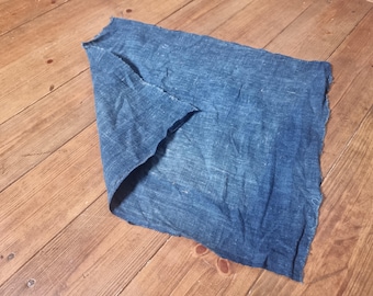 May include: A piece of blue fabric with a raw edge, lying on a wooden surface. The fabric has a textured appearance and a slightly crumpled look. The color is a deep, saturated blue.
