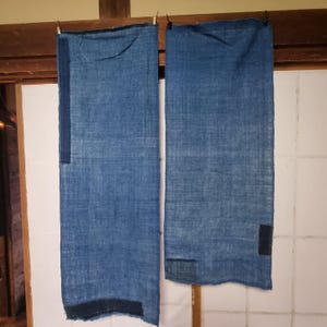 Puede incluir: Dos paneles de tela azul índigo colgados de una viga de madera. Cada panel tiene parches azules más oscuros cosidos, dando un aspecto texturizado. Los paneles están suspendidos por pinzas, lo que sugiere una calidad artesanal o hecha a mano. El fondo es una pared de color claro.