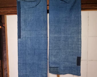 May include: Two indigo blue fabric panels hanging from a wooden beam. Each panel has darker blue patches sewn on, with a textured appearance. The panels are suspended by clothespins, suggesting a handmade or artisanal quality. The background is a light-colored wall.