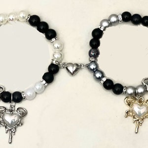 Puede incluir: Dos pulseras de cuentas conectadas por un dije en forma de corazón. Una pulsera tiene cuentas negras y blancas, la otra tiene cuentas negras y grises. Ambas pulseras tienen un dije en forma de corazón con un diseño de goteo. El dije en forma de corazón de la pulsera negra y blanca es plateado, y el dije en forma de corazón de la pulsera negra y gris es dorado.