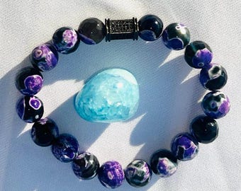 Puede incluir: Un brazalete de cuentas de ágata negro y morado con un acento de cuentas de metal negro. El brazalete está sobre una superficie blanca junto a una piedra de ágata azul claro.