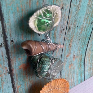 May include: A seashell mobile with a variety of shells, including a brown shell, a green and blue shell, and a white shell with a green glass bead inside. The shells are wired together and hang from a wire.