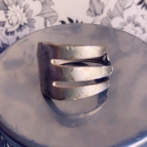 May include: A silver ring made from a repurposed fork. The ring has three prongs and a rustic, worn look.