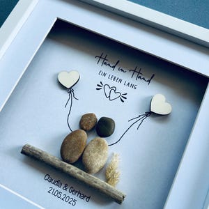 May include: A framed artwork featuring a pebble art design. Two heart-shaped wooden cutouts are connected by drawn lines to a pair of pebbles. The text "Hand in Hand EIN LEBEN LANG" is written above the pebbles. The names "Claudia & Gerhard" and the date "21.05.2025" are below.