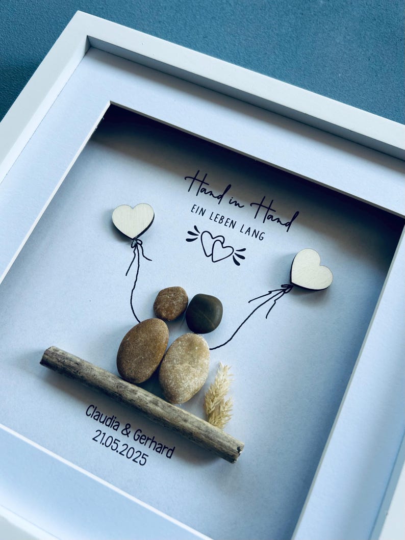 May include: A framed artwork featuring a pebble art design. Two heart-shaped wooden cutouts are connected by drawn lines to a pair of pebbles. The text "Hand in Hand EIN LEBEN LANG" is written above the pebbles. The names "Claudia & Gerhard" and the date "21.05.2025" are below.