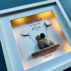 May include: A framed artwork with a white border, featuring a pebble art scene. Two heart-shaped wooden cutouts are attached to the top with drawn lines. The text "Hand in Hand EIN LEBEN LANG" is written at the top. The names "Claudia & Gerhard" and the date "21.05.2025" are at the bottom.
