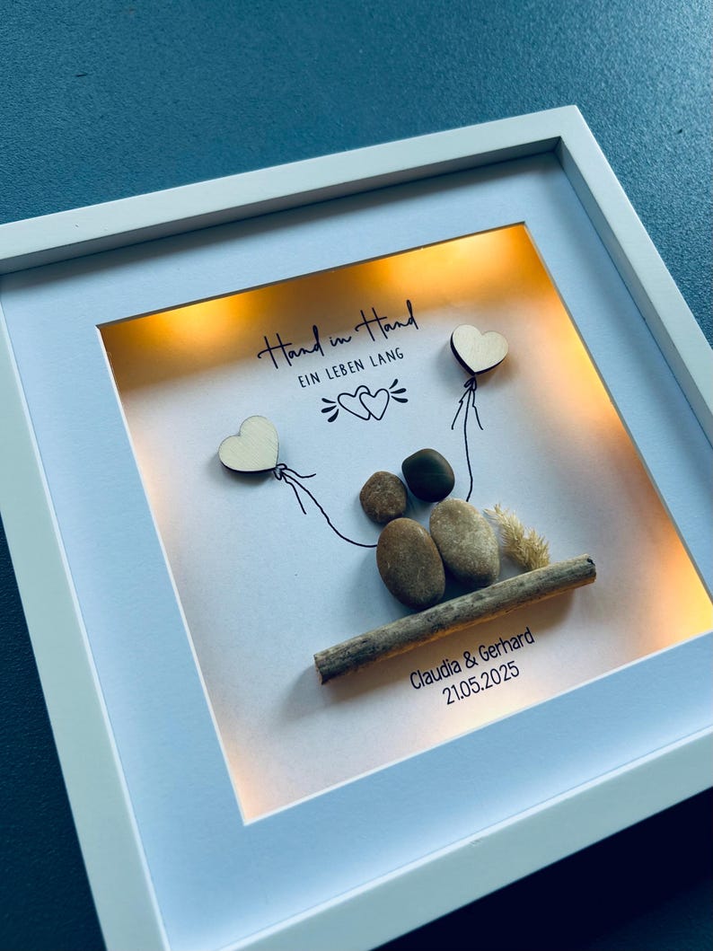 May include: A framed artwork with a white border, featuring a pebble art scene. Two heart-shaped wooden cutouts are attached to the top with drawn lines. The text "Hand in Hand EIN LEBEN LANG" is written at the top. The names "Claudia & Gerhard" and the date "21.05.2025" are at the bottom.