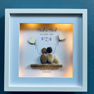 May include: A framed artwork with a white border, featuring a pebble art scene. The art includes two pebble figures, a wooden branch, and the text "Hand in Hand EIN LEBEN LANG". Two wooden heart shapes are held by the figures. The names "Claudia & Gerhard" and the date "21.05.2025" are also included.
