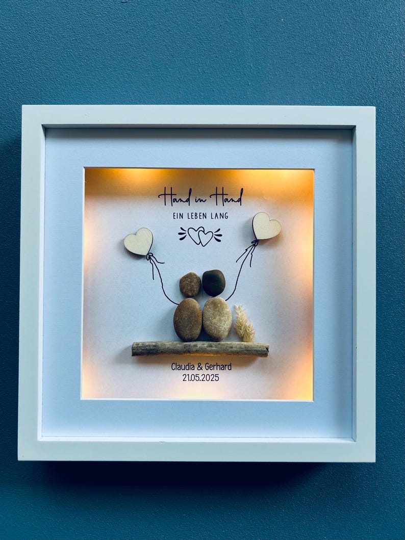 May include: A framed artwork with a white border, featuring a pebble art scene. The art includes two pebble figures, a wooden branch, and the text "Hand in Hand EIN LEBEN LANG". Two wooden heart shapes are held by the figures. The names "Claudia & Gerhard" and the date "21.05.2025" are also included.